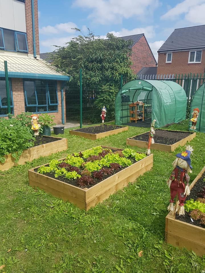 The Tastebuds community garden: raised vegetable beds, painted scarecrows and a green polytunnel beside a brick building