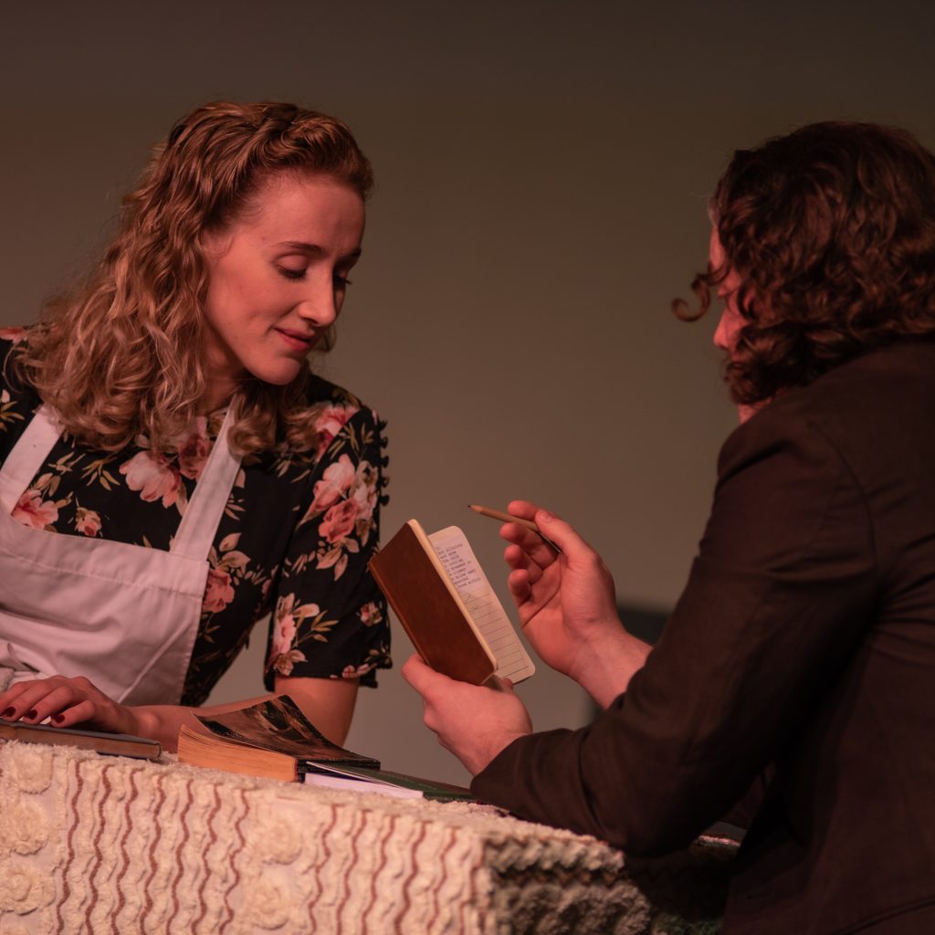 Two actors performing a scene from Beyond the Pier, one in an apron behind a counter and one holding a small notebook