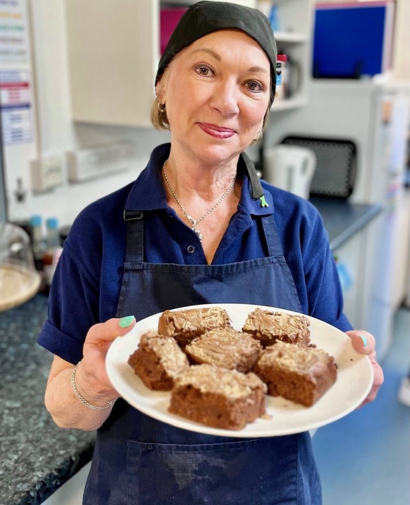 A Café Camaraderie team member in a navy polo and apron holding a plate of freshly baked tray bakes