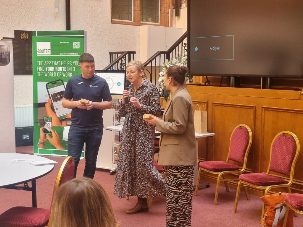 Speakers addressing guests at Social Enterprise Day in a wood-panelled hall, with Routes app banners behind them