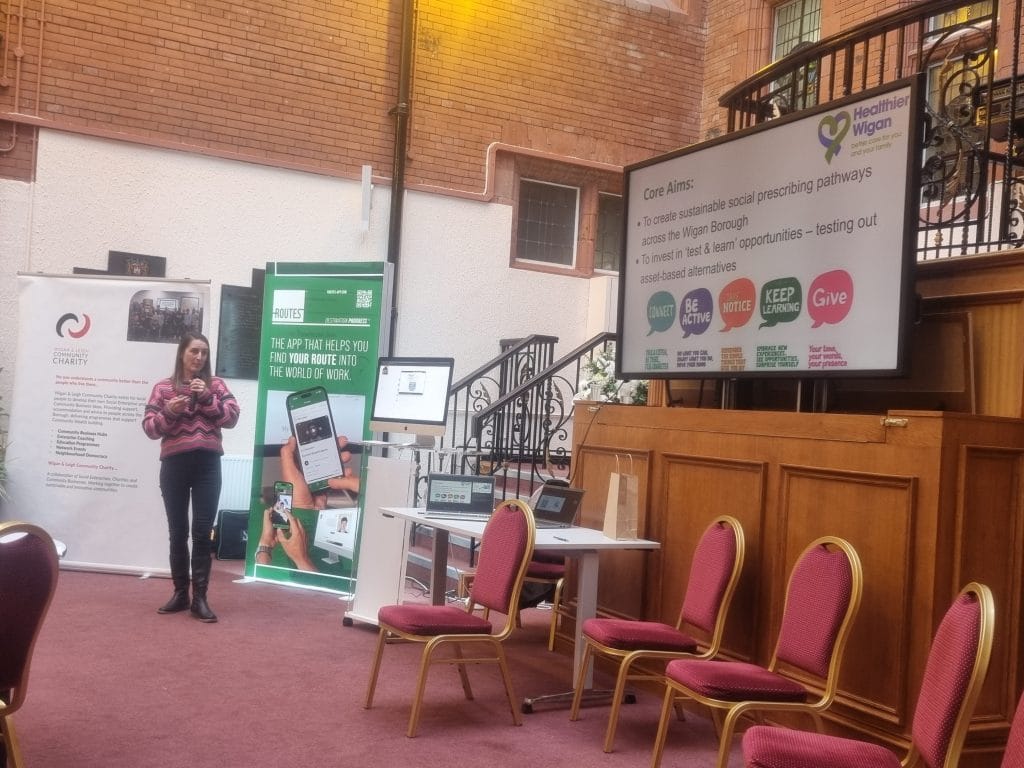 A speaker presenting the core aims of Wigan's social prescribing pathways, beside a Healthier Wigan slide