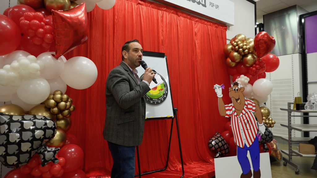 A host with a microphone presenting beside a flip chart at the Proper Good pop-up, surrounded by balloons