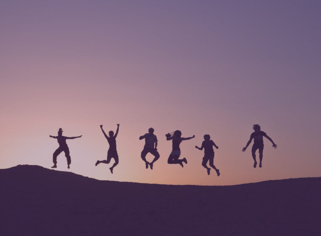 Silhouettes of six people jumping on a hilltop against a purple sunset