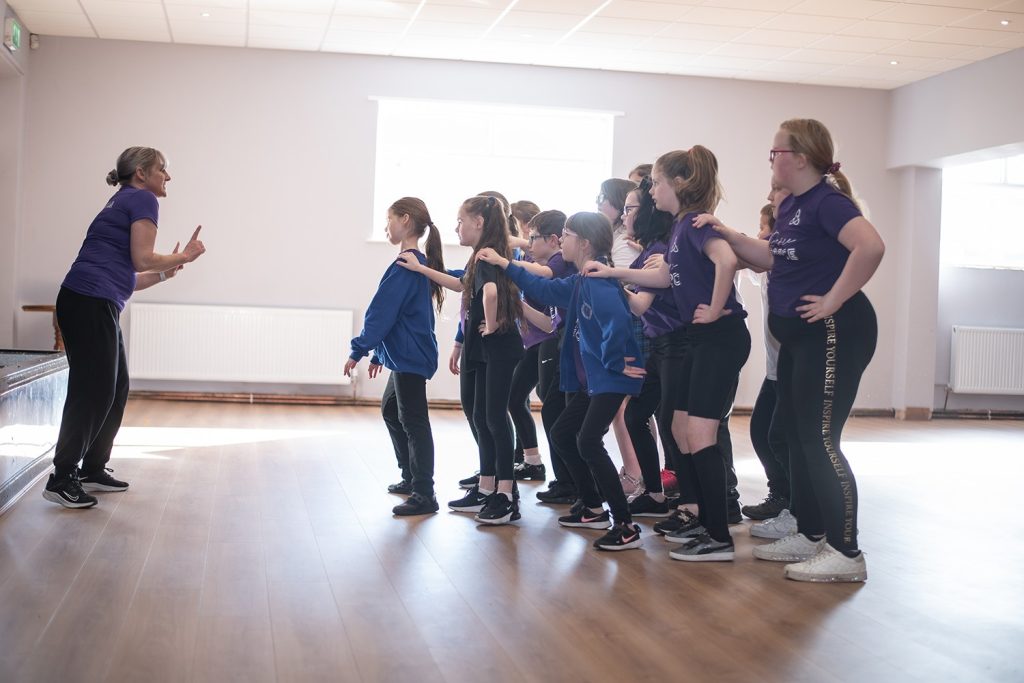 Young dancers lined up with hands on each other's shoulders as their teacher leads the class