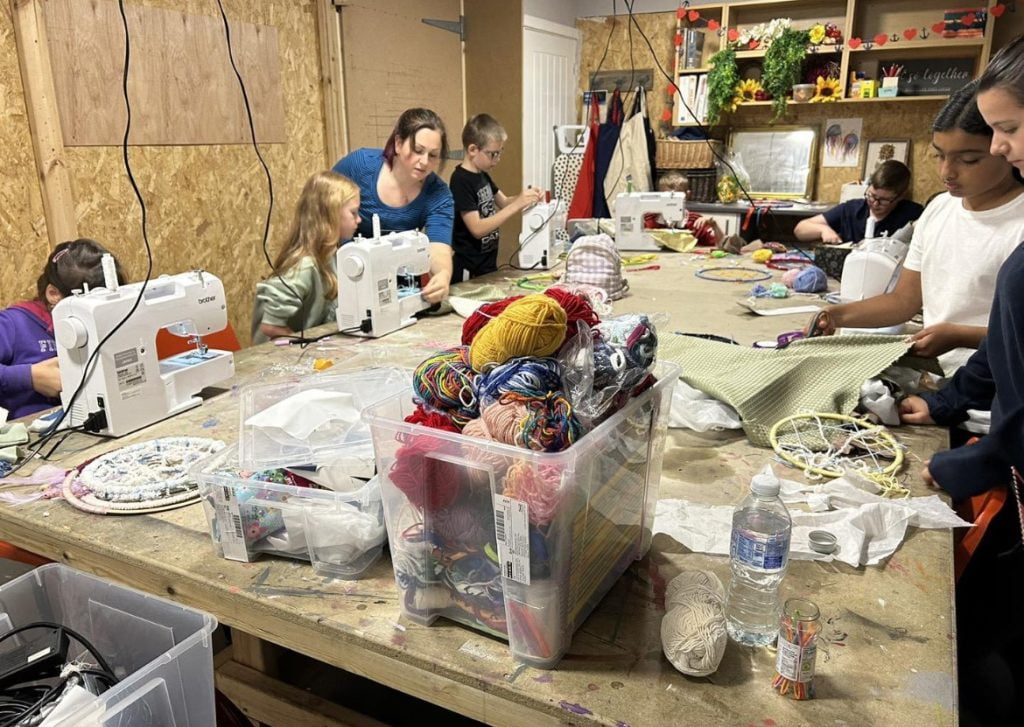 Young people working at sewing machines around a craft table stacked with yarn and fabric