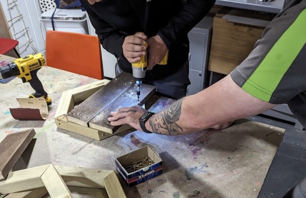 Close-up of hands driving a screw into timber with a cordless drill while an instructor steadies the wood